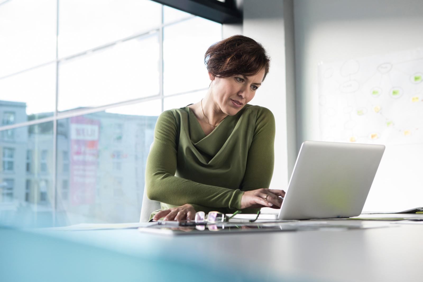 Woman looking at laptop in a room with a glass window behind her.