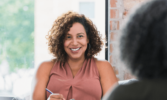 Woman smiling warmly while holding a pen and having a discussion with a person across from her.