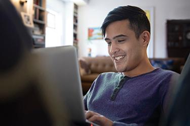 Male student sitting in his living room at home. He is paying online bills on his laptop.